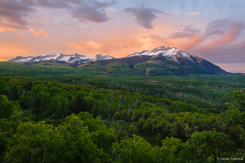 The clouds behind the Beckwith Mountains glow with the light of the rising sun beyond a valley filled with vibrant green aspens outside of Crested Butte, Colorado.