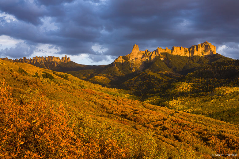 The setting sun breaks through clouds and bathes Chimney Rock, Courthouse Mountain, and the valley filled with autumn colors below with golden light outside of Ridgway, Colorado.