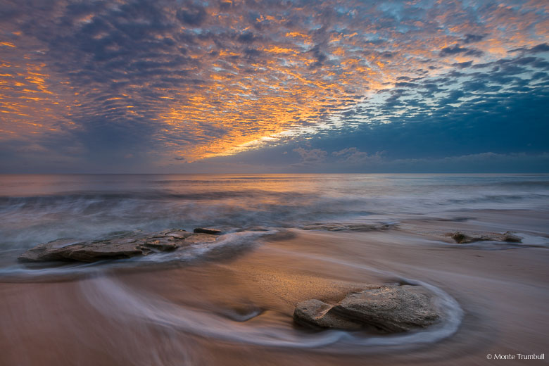 The rising sun illuminates the breaking cloud cover over a rocky beach in Washington Oaks Gardens State Park, Florida.