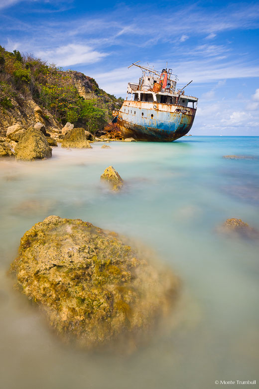 The rusting remains of a grounded ship rests along the shore surrounded by the colorful waters of Road Bay in Anguilla, BWI.