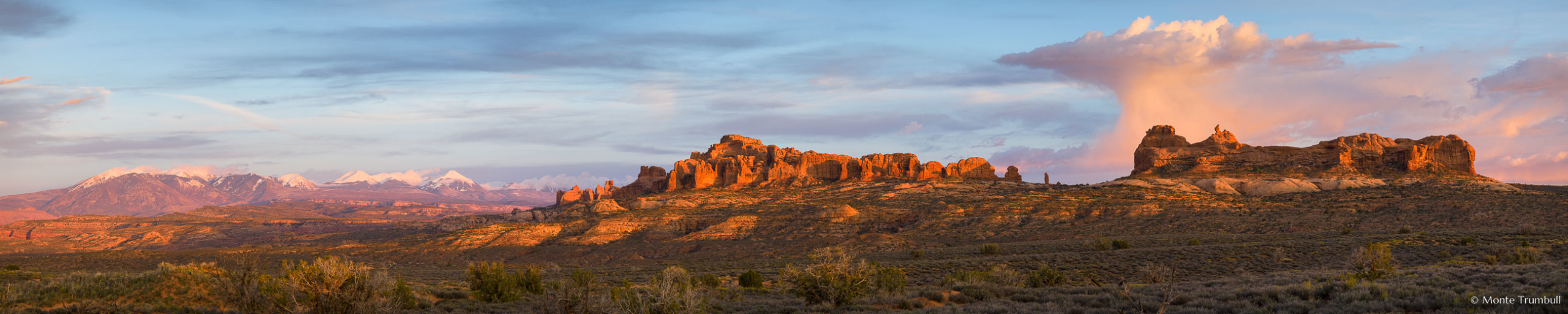 The setting sun casts late day shadows on the Garden of Eden rock formations and illuminates a towering cloud formation above it as well as the distant snow-covered La Sal Mountains in Arches National Park, Utah.