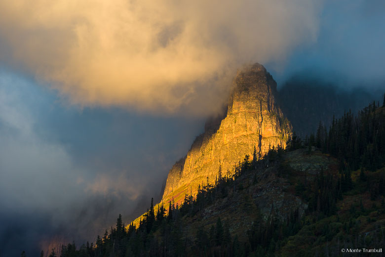 A shaft of light streams through swirling clouds and illuminates the side of Pumpelly Pillar at sunrise in Glacier National Park, Montana.