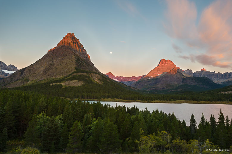 The first rays of sunlight strike the tops of Grinnell Point and Swiftcurrent Mountain as the full moon sets between them at Swiftcurrent Lake in Glacier National Park in Montana.