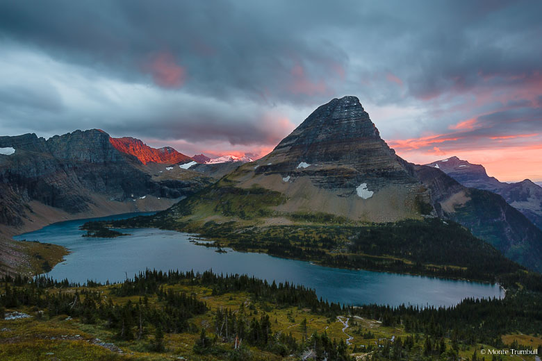 The clouds turn pink and a shaft of light paints the mountains red behind Bearhat Mountain and Hidden Lake as the sun sets high in Glacier National Park, Montana.