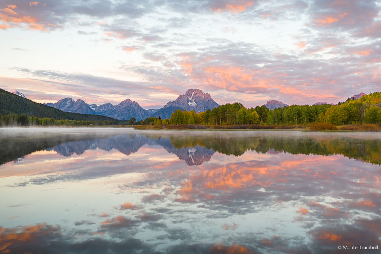 Mount Moran and a sky filled with glowing pink clouds at sunrise are reflected in the calm waters of the Snake River at Oxbow Bend in Grand Teton National Park, Wyoming.