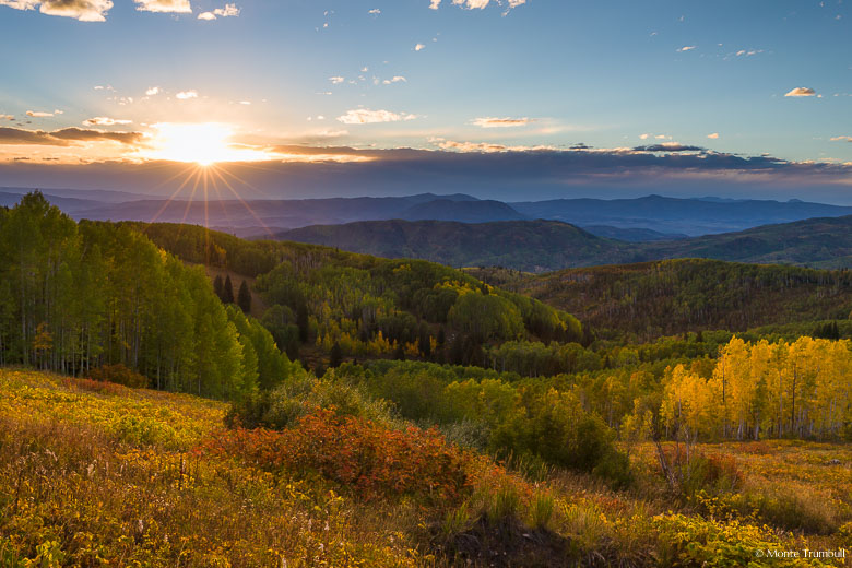 The sun sets through distant clouds beyond a golden hillside and a valley dotted with vibrant fall color outside of Steamboat Springs, Colorado.