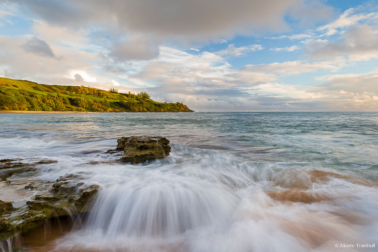 Waves rush into the rocky shoreline in Moloaa Bay as the early morning light illuminates the lush green hillside in the distance on the northeast coast of Kauai, Hawaii.