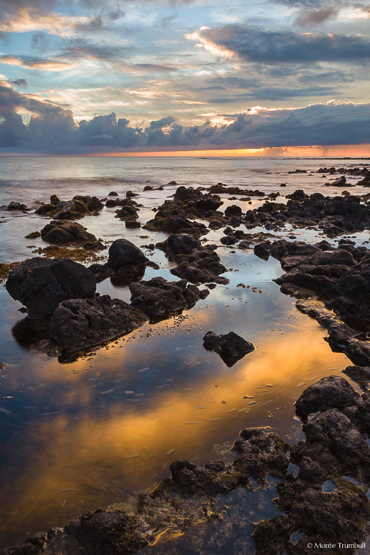 The setting sun peeks out from behind a cloud bank and reflects in tide pools along the rocky shore at Poipu Beach Park in Kauai, Hawaii.