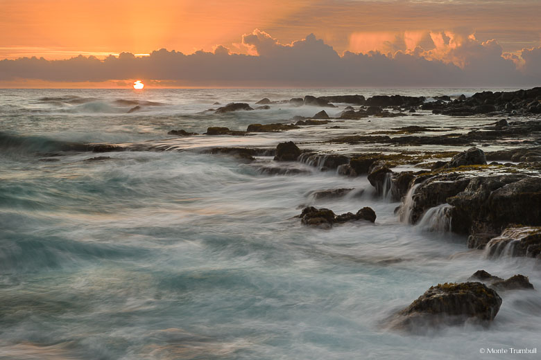 The orange ball of the sun radiates shafts of light skyward through the clouds as it rises over the horizon off a rocky shoreline pounded by waves on the east coast of Kauai, Hawaii.