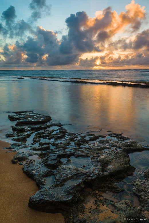 The sun rises behind a bank of clouds, filling the sky with golden light and reflecting in a pool along a rocky shoreline on Waipouli Beach in Kauai, Hawaii.