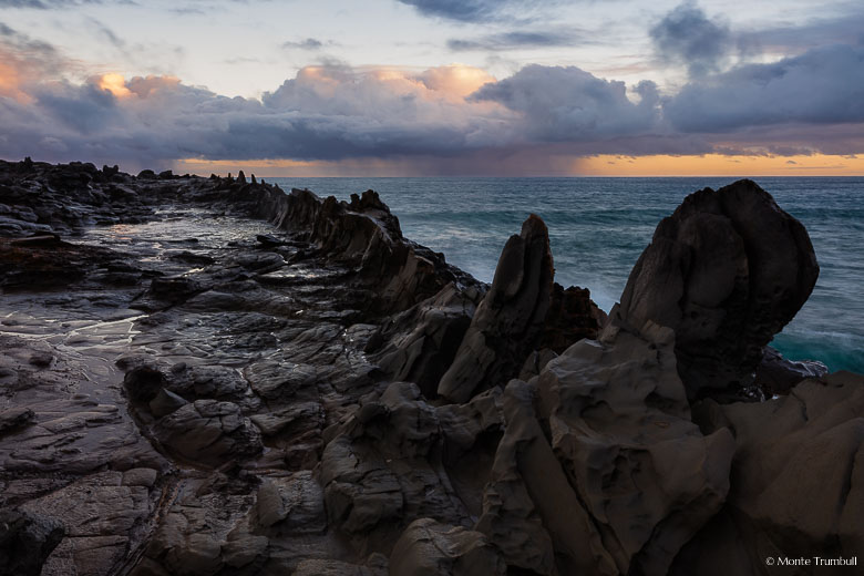 Stormy clouds and a rain squall over the ocean are illuminated by early morning light beyond the rocky Dragon's Teeth formation on the northwest coast of Maui, Hawaii.