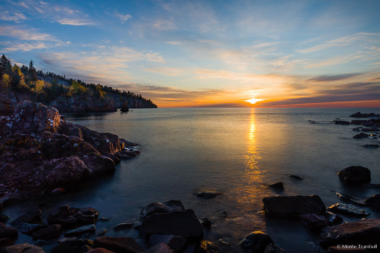 The sun peeks through the clouds as it rises above Lake Superior, shining light on Shovel Point and a rocky shoreline in Tettegouche State Park along the North Shore of Minnesota.