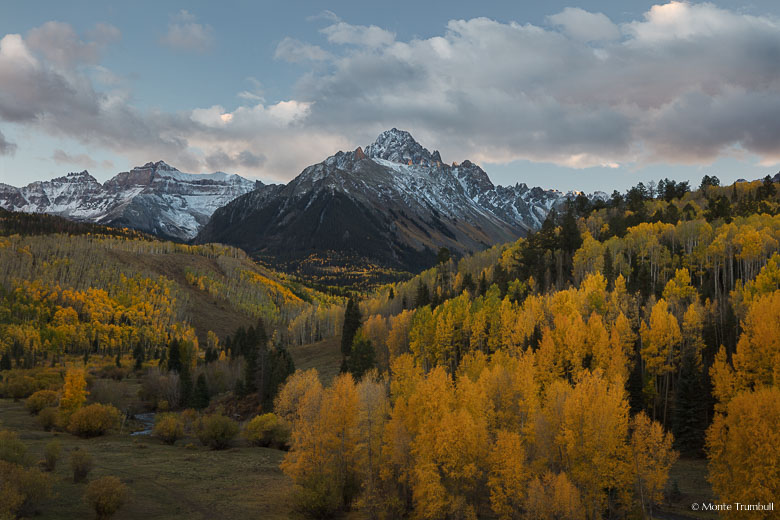 A few rays from the setting sun shine through glowing clouds and illuminate the side of Mount Sneffels above a valley filled with golden aspen trees in the Uncompahgre National Forest outside of Ridgway, Colorado.