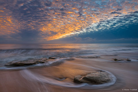 The rising sun illuminates the breaking cloud cover over a rocky beach in Washington Oaks Gardens State Park, Florida.