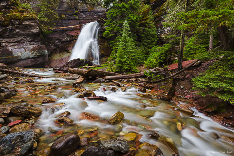 Baring Creek gently flows over Baring Falls and glides down a streambed sprinkled with red rocks on its way down to Saint Mary Lake in Glacier National Park in Montana.